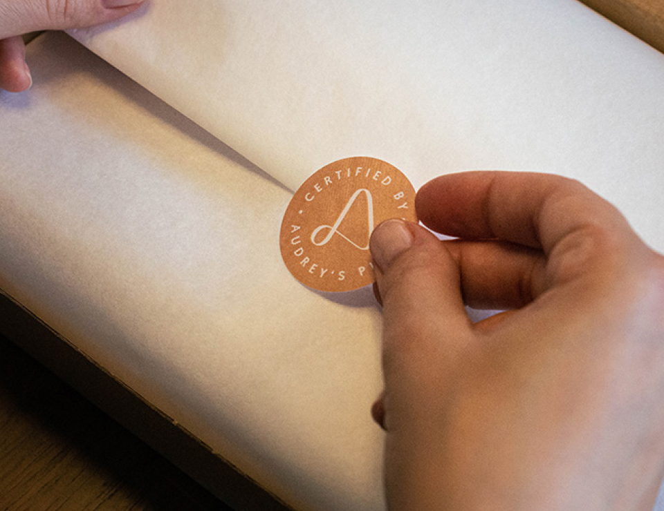 Hand, die Aufkleber mit Signet "Audrey's Place" auf Verpackung aufklebt