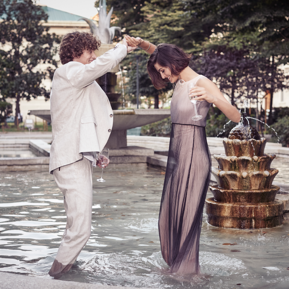 Mann im Anzug und Frau im Abendkleid tanzen lachend barfuß in einem Brunnen, beide mit Weingläsern in der Hand.