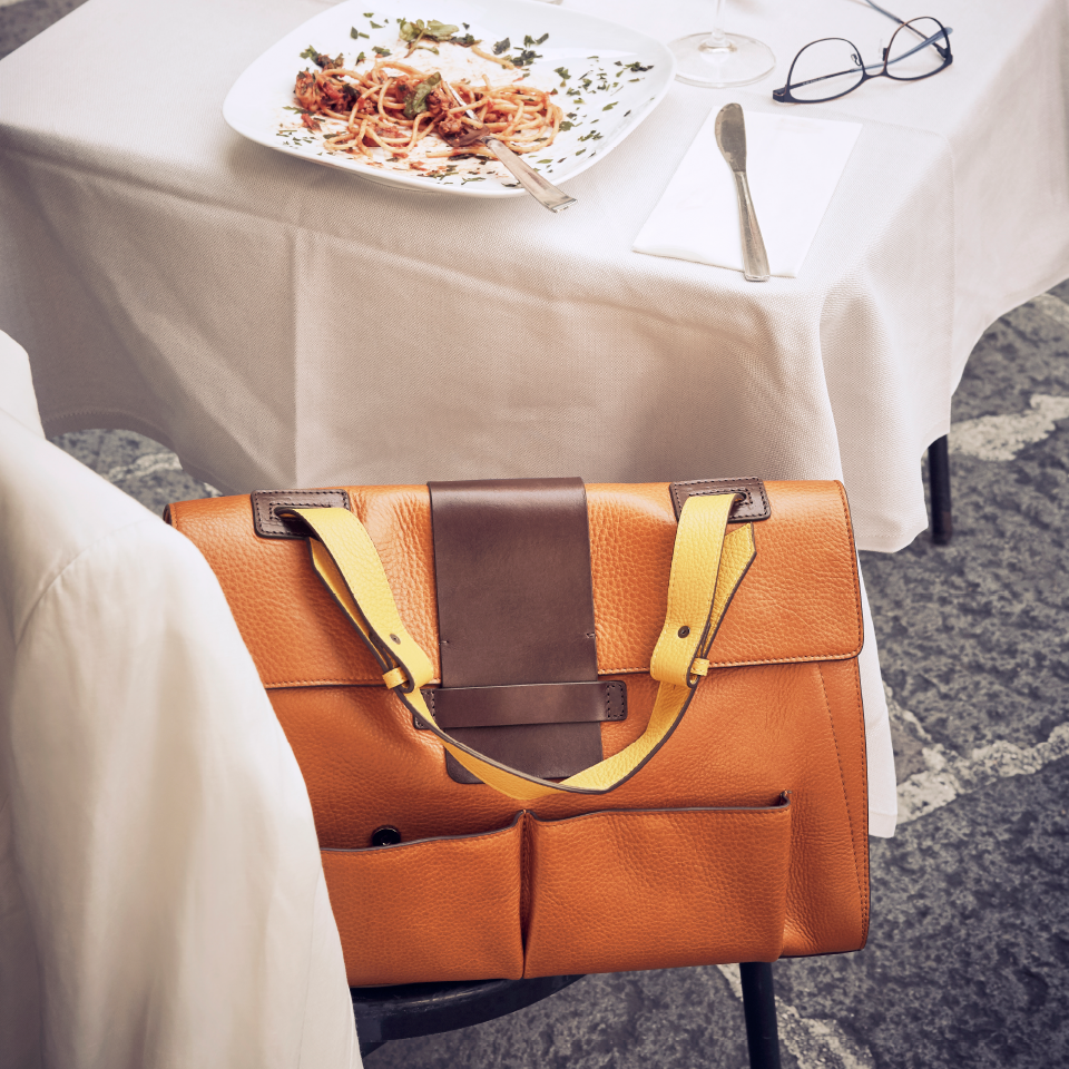 Eine hochwertige Lederhandtasche steht in in einem Restaurant auf dem Stuhl. Auf dem Tisch eine Brille und ein Teller Spaghetti.