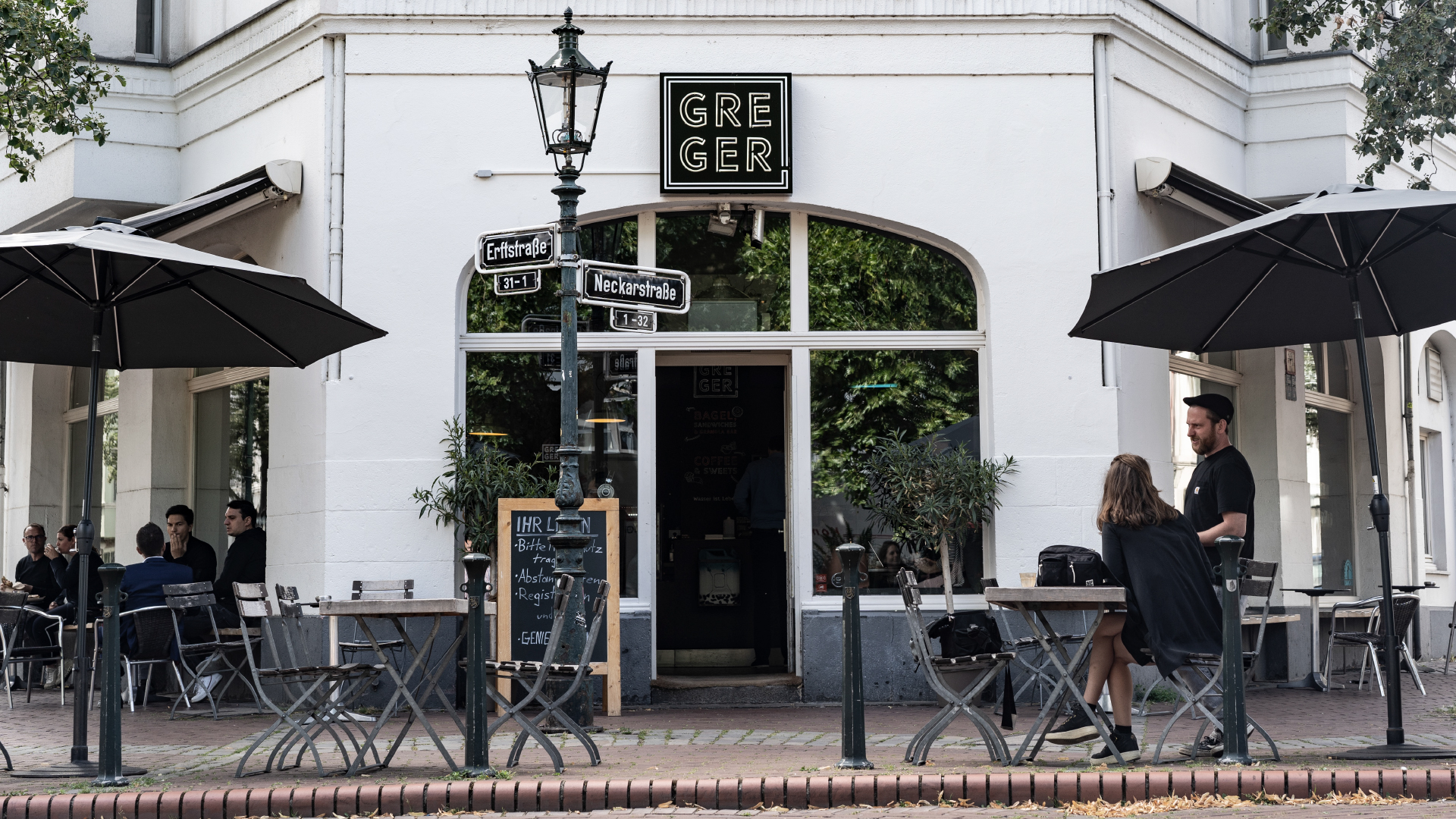 Außenansicht des Cafés GREGER mit Tischen, Stühlen und Sonnenschirmen auf der Terrasse, Gästen und dem Inhaber im Gespräch.