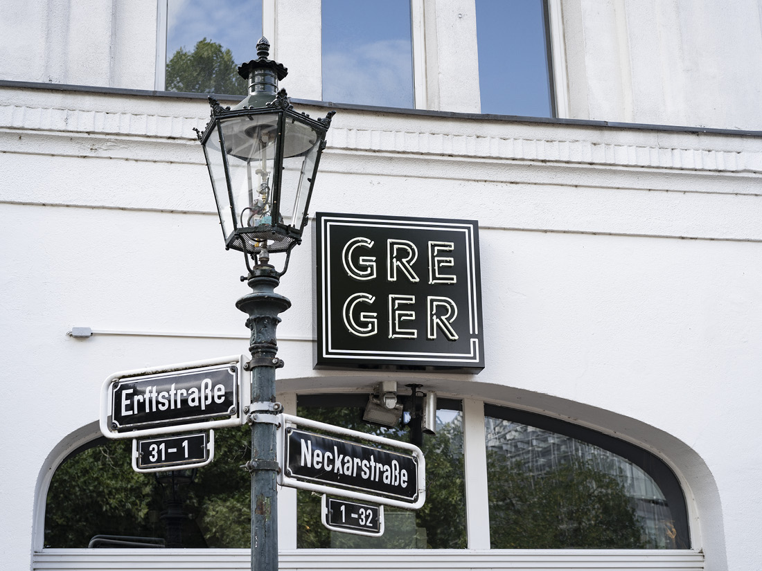 Leuchtschild „GREGER“ an Gebäudefassade mit Straßenschildern und Straßenlaterne.