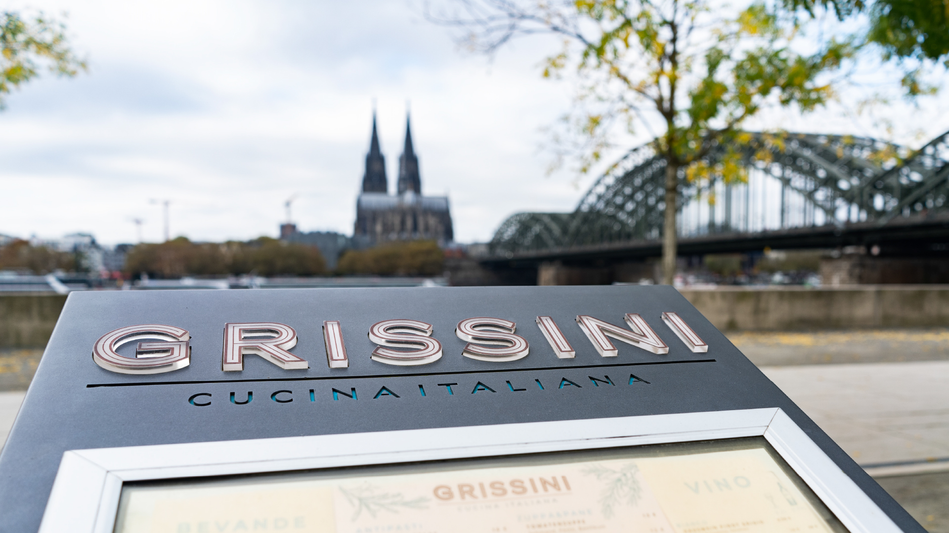 Restaurantschild „Grissini Cucina Italiana“ im Vordergrund mit Kölner Dom und Hohenzollernbrücke im Hintergrund.