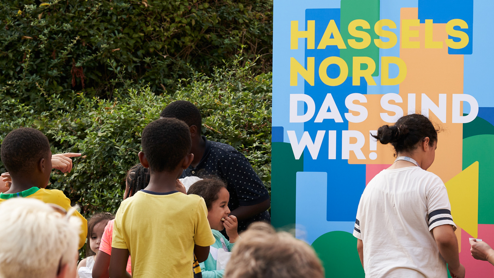 Kinder und Erwachsene stehen vor einem großen bunten Banner mit der Aufschrift „Hassels Nord – Das sind wir!“.