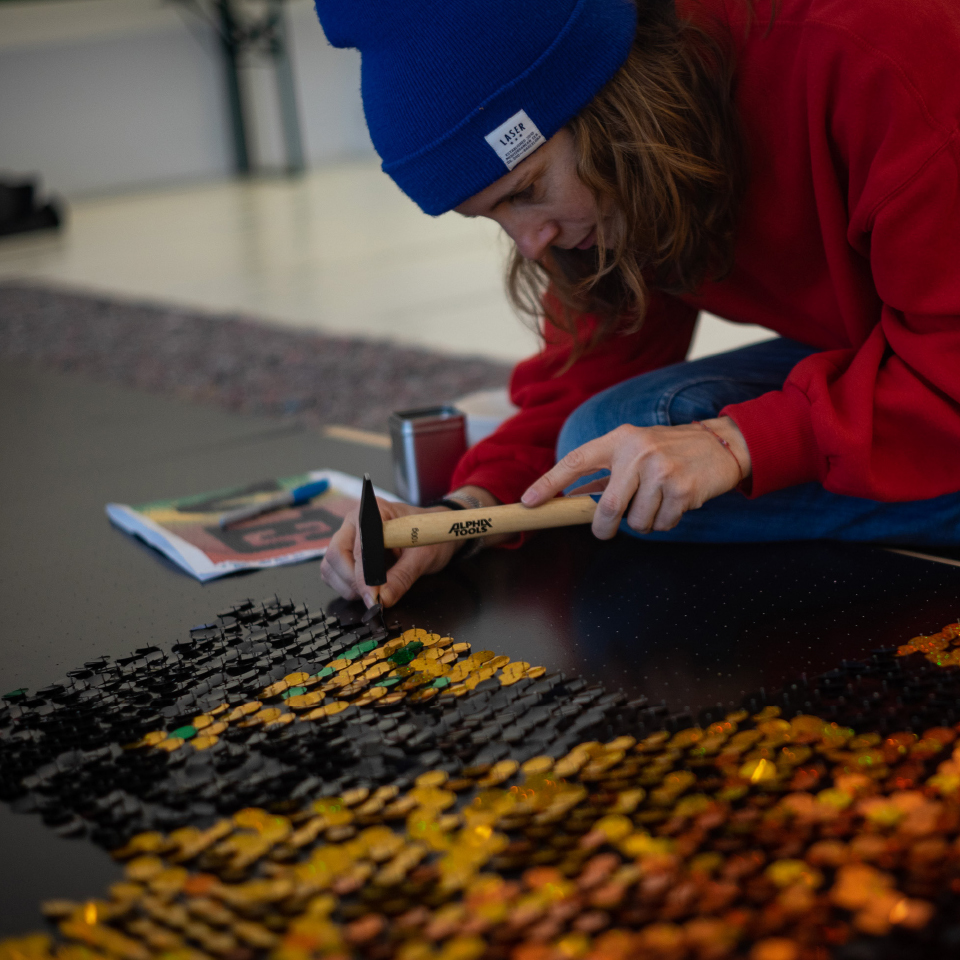 Person mit blauer Mütze und rotem Pullover hämmert bunte Pailletten in ein Kunstwerk ein.