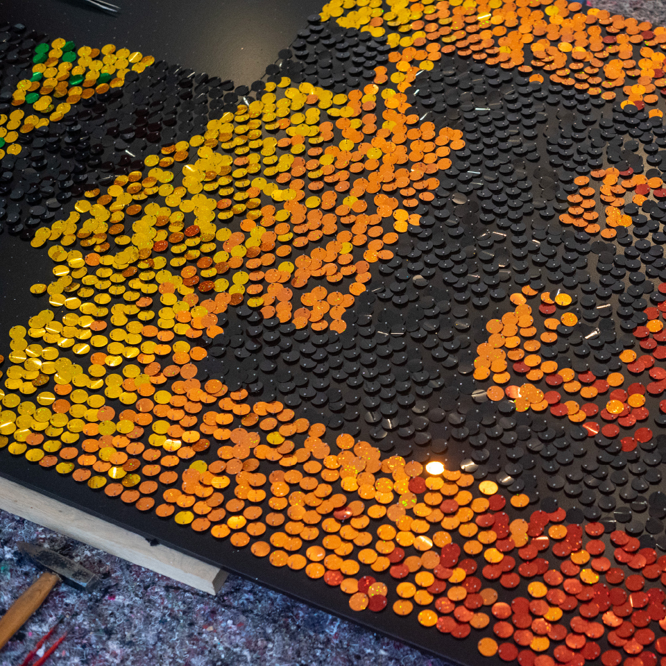 Detailaufnahme eines Kunstwerkes mit bunten Pailletten in Gelb-, Orange- und Rottönen auf schwarzem Hintergrund.