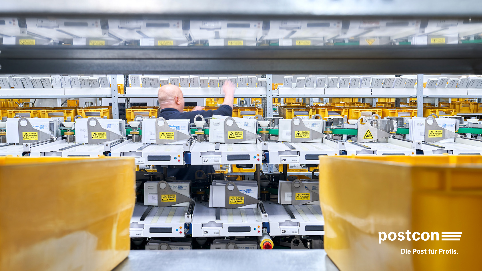 Sortieranlage bei Postcon mit gelben Postkisten und einem Mitarbeitenden im Hintergrund.
