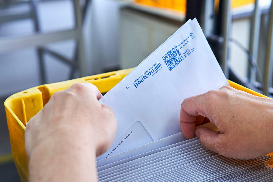 Hände sortieren einen Brief mit Postcon-Logo in eine gelbe Postkiste.