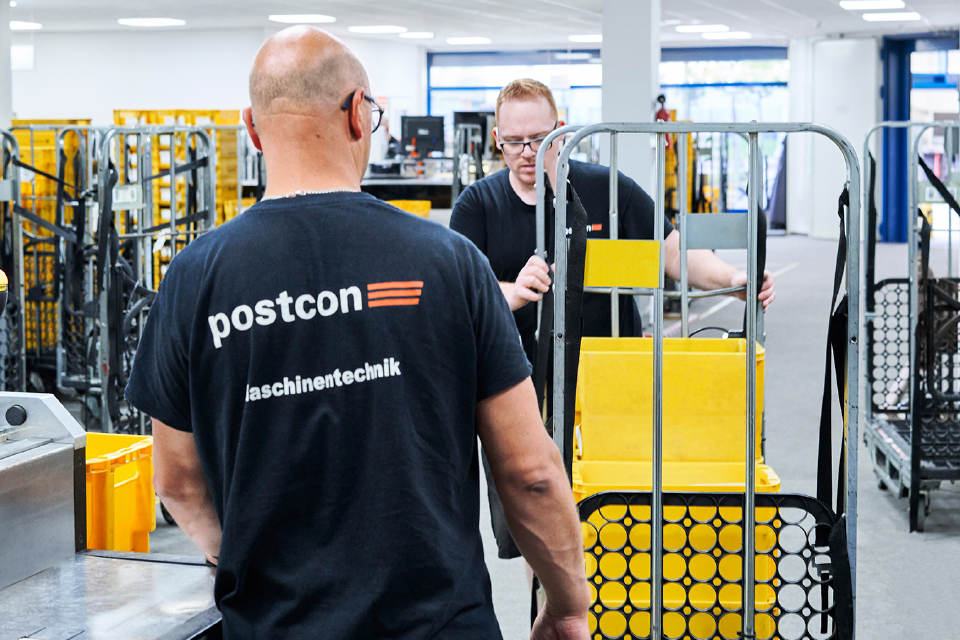 Zwei Postcon-Mitarbeiter bewegen gelbe Postbehälter auf einem Rollwagen.