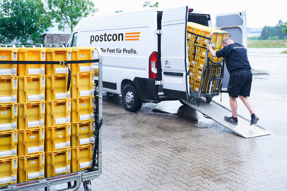 Postcon-Mitarbeiter schiebt gelbe Postbehälter auf einem Rollwagen in ein Lieferfahrzeug.