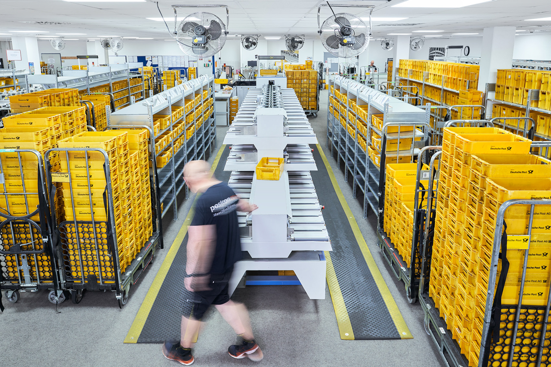Blick in eine Postcon-Sortierhalle mit Regalen und Stapeln gelber Postkisten.