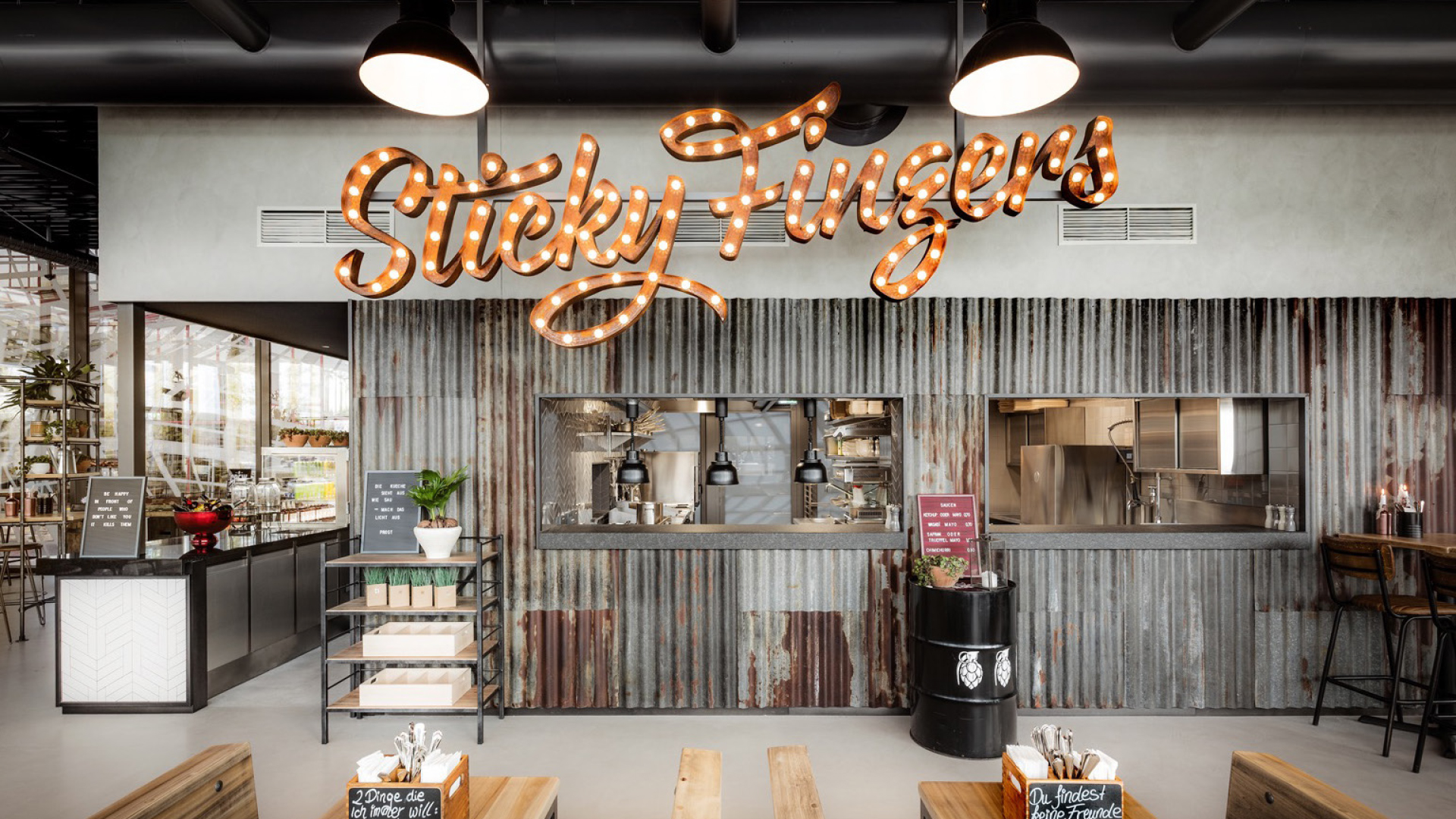 Restaurantinnenraum mit offener Küche, Wellblechverkleidung und beleuchtetem Schriftzug „Sticky Fingers“.