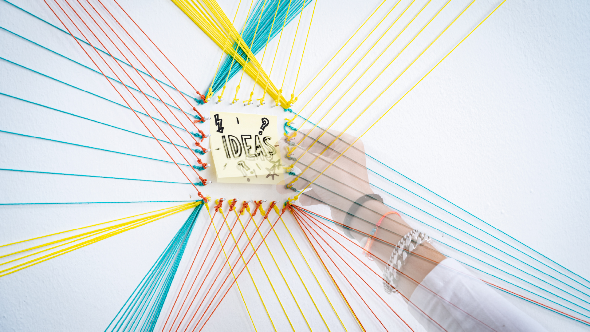 Hand platziert Post-it mit der Aufschrift „IDEAS“ in einem Netzwerk bunter Fäden an einer Wand.