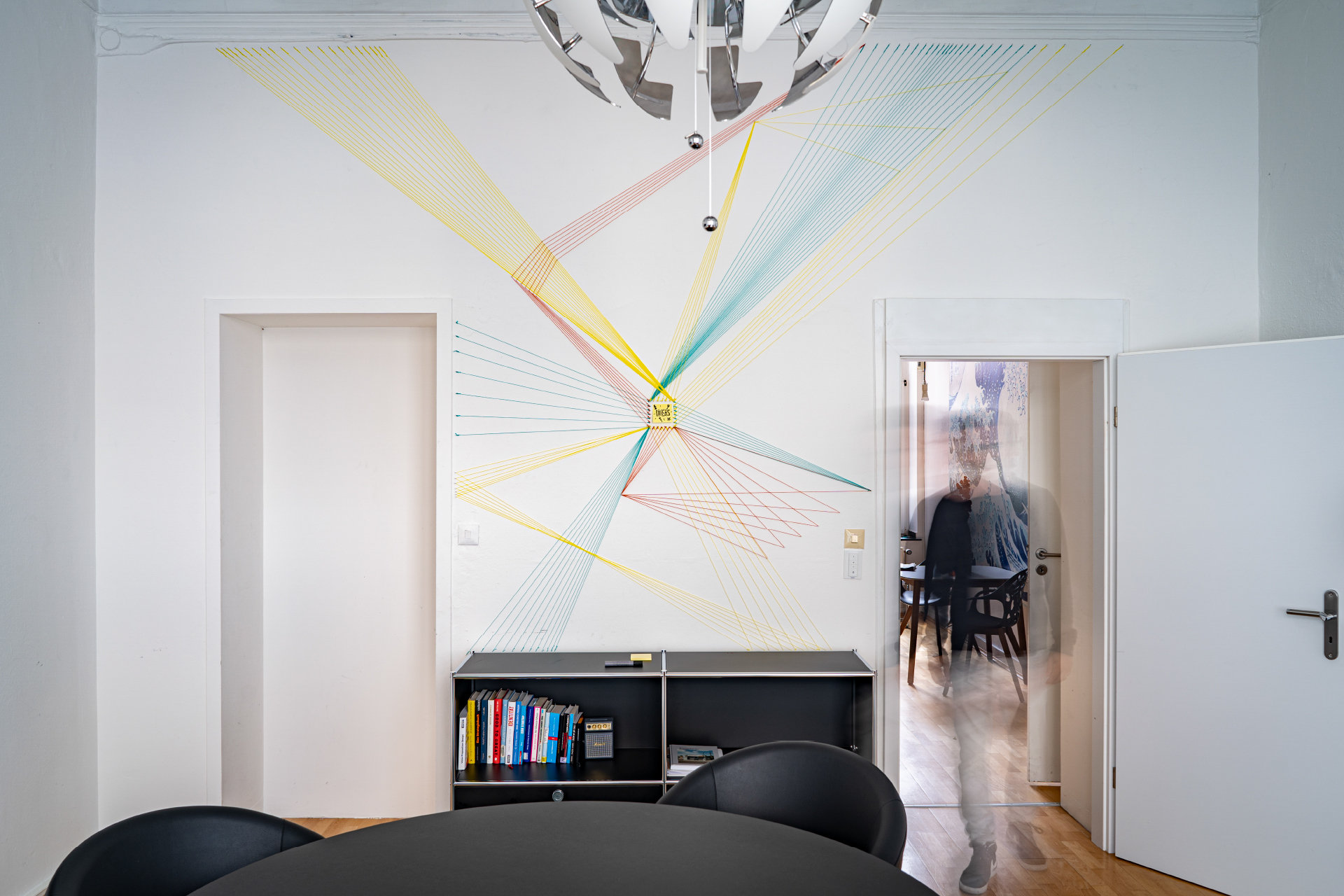Besprechungsraum mit Wandkunst aus bunten Fäden, Bücherregal und geöffneter Tür.