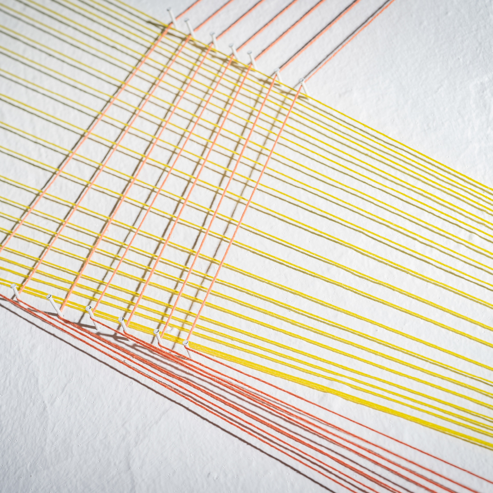 Detailaufnahme einer Wandinstallation mit gespannten gelben und roten Fäden, die über weiße Wandnägel verlaufen.