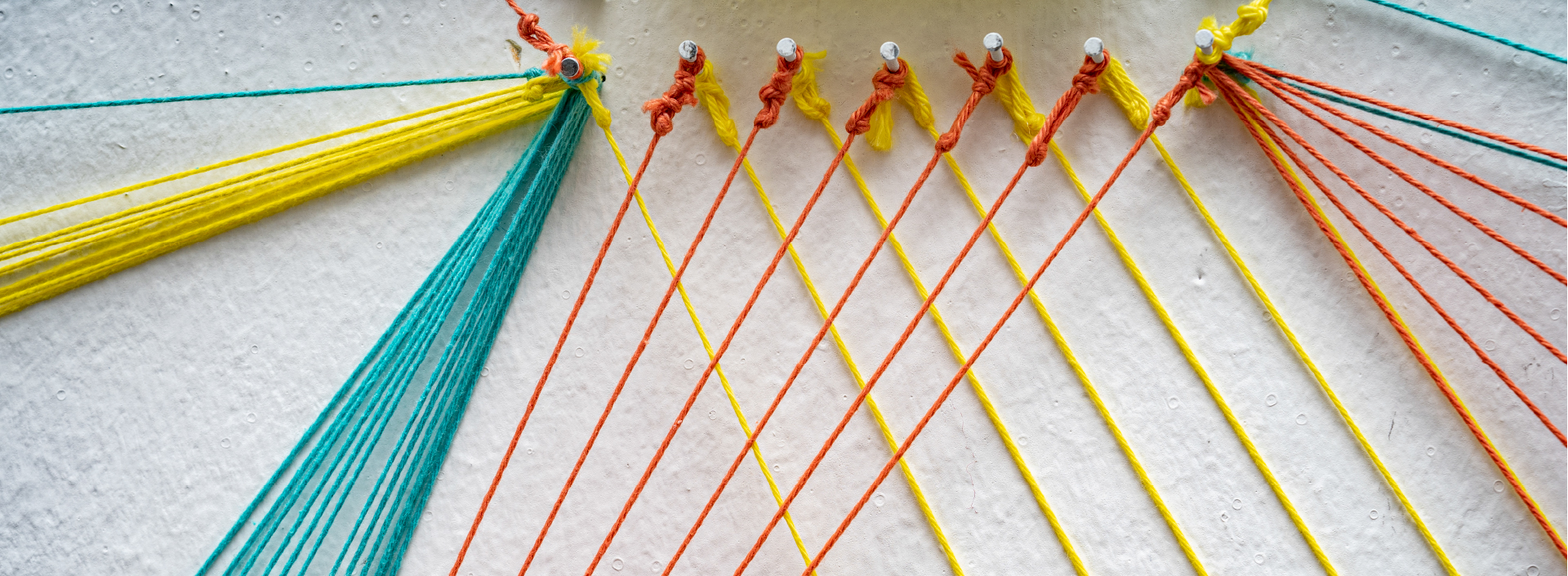 Detailaufnahme einer Wandinstallation mit gespannten gelben, roten und grünen Fäden, die über weiße Wandnägel verlaufen.