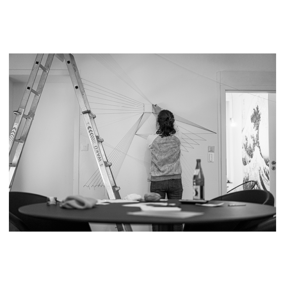 Schwarz-Weiß-Foto: Person arbeitet an einer Wandinstallation mit gespannten Fäden, daneben steht eine Leiter, im Vordergrund ein Tisch mit Utensilien.
